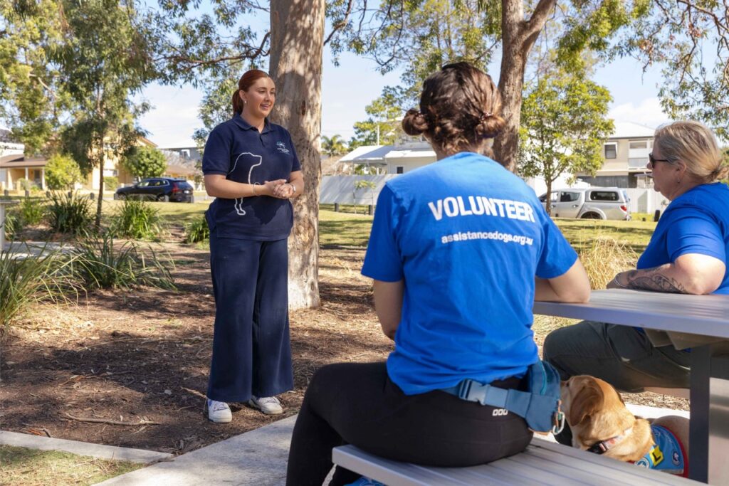 Georgia, an instructor with Assistance Dogs Australia, speaking with two seated volunteers, all from Assistance Dogs Australia.
