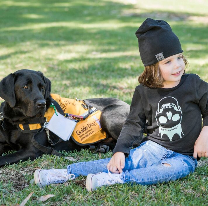 Zena, the Autism Assistance Dog, a black Labrador in her orange working Guide Dogs Vest sitting with a young boy in the grass.